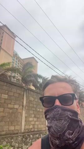 A person wearing sunglasses and a patterned face covering is shown in a selfie-style video. They are standing outdoors in front of a light pink building with multiple windows and a blue-domed roof. A tall, concrete utility pole with wires runs through the frame. Behind the person is a brick wall. The sky is overcast.