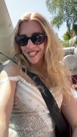 The video shows a young woman with blonde hair and sunglasses in the passenger seat of a convertible. She is smiling and posing for the camera, occasionally making a peace sign with her fingers. The background shows a street with other cars and trees, suggesting the car is in motion. The woman is wearing a white lace top and a seatbelt.
