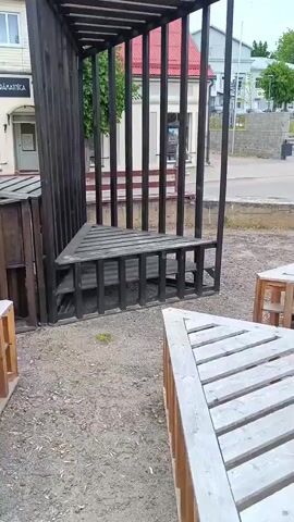The video presents an outdoor scene in Aizpute, Latvia, featuring several contemporary wooden structures. The primary subject is a tall, dark-colored, open-sided structure resembling a cage or minimalist pergola, constructed from vertical and horizontal wooden beams. Inside, a triangular, slatted wooden base serves as a seat or platform. In the foreground, two other triangular wooden objects, possibly benches or low tables, feature light-colored slatted tops and darker wooden sides.

As the camera pans to the right, a rectangular wooden planter box filled with vibrant yellow flowers, likely marigolds, becomes visible behind one of the triangular structures. The background reveals two-story buildings; one has a striking red roof and light-colored walls, while the adjacent building displays light-colored siding. On the facade of the latter, a sign reads "ZVAIGZNES GRĀMATNĪCA," identifying it as a bookstore in Latvian.

The setting appears to be a public square or park area, with a gravelly ground surface in the immediate vicinity of the wooden installations, transitioning to a paved area further back. Trees and other greenery can be seen in the distance. The lighting suggests an overcast daytime, but no specific weather conditions or activities are discernible, and no people are present.