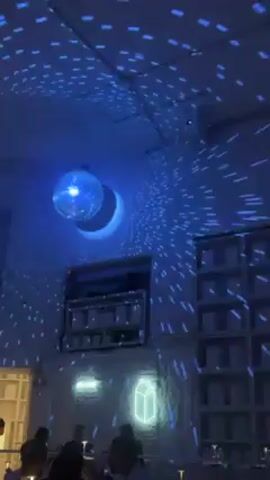 Here's a description of the video:

The video shows the interior of a bar or restaurant. A disco ball hangs from the ceiling, casting blue light spots across the walls and ceiling. People are sitting at tables, and shelves stocked with items line one wall. A neon light in the shape of a milk carton is also visible.