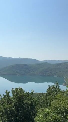 The video shows a scenic view of a lake surrounded by lush green hills and mountains. The sky is clear and blue, and the water in the lake is calm and reflects the surrounding landscape. In the foreground, there are trees with dense foliage. The camera pans slowly across the landscape, revealing islands with trees scattered throughout the lake. The overall impression is one of tranquility and natural beauty.