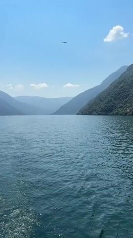 The video opens with a wide shot of a tranquil lake surrounded by lush green mountains under a clear blue sky. A single bird is visible soaring high above in the distance. As the video progresses, the camera pans to the right, revealing houses nestled on the slopes of the mountains. The water in the foreground exhibits gentle ripples, indicating the presence of a boat or a slight breeze. The houses appear to be traditional in design, with terracotta roofs and white or pastel-colored walls, interspersed with dense foliage. The scene is peaceful and idyllic, capturing the natural beauty of the mountainous lakeside environment.