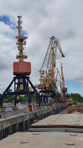 The video shows a port with three large, fixed cranes lined up along a concrete dock. The cranes are painted in various shades of beige, yellow, and dark brown.  The dock itself is somewhat cracked and worn, with large concrete blocks lining the edge.  A man wearing a red hard hat and work clothes walks along the dock.  The video is shot from the deck of a boat or barge, with the side of the vessel visible in the foreground. The sky is cloudy with patches of blue.
