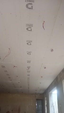 The video shows the interior of a room under construction. The walls are unfinished, with exposed concrete and markings. The ceiling is covered with white panels bearing the logo "DONICY". Wires hang down from the ceiling in several places. There is a doorway visible in the background, and a window providing natural light. The room appears to be empty of furniture and other fixtures. The camera moves slightly, panning across the room, focusing primarily on the ceiling and upper parts of the walls.