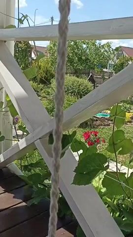 The media file shows a close-up view of a white wooden railing with a thick rope hanging in front of it. Behind the railing, a lush green garden is visible, with various plants, flowers, and trees. In the background, there are houses with red roofs, and a wooden fence. The sky is blue with white clouds, suggesting a bright and sunny day. The scene appears to be set in a residential area, possibly a backyard or a balcony overlooking a garden. The overall impression is one of a peaceful and serene outdoor setting. The time of day is likely daytime, given the bright sunlight. No people are visible in the frame.