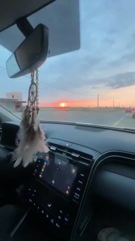 The video shows a car interior during a sunset. The view is from the driver's seat, looking out at a highway. The sun is setting in the distance, casting a warm orange glow across the sky. A dreamcatcher hangs from the rearview mirror, partially obscuring the view. The car's dashboard is visible, including the navigation screen and various buttons. Other cars are on the road as well.