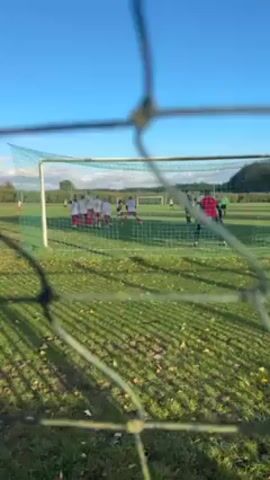 The video, shot from inside a soccer goal, shows a soccer match in progress on a green field under a clear blue sky. Several players, mostly wearing red and white, are visible around the opposing goal. A goalkeeper, wearing green, is on the ground after what appears to be a save. The camera's viewpoint is through the mesh of the goal, which is slightly blurred. The background includes trees and vegetation in the distance.