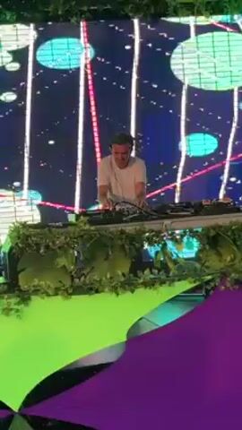 A person is DJing on a stage with a large screen behind them displaying abstract designs. The stage is decorated with greenery. A photographer in a cap, t-shirt, and shorts is taking photos of the event from the ground. The ground is decorated with green and purple designs. The video is taken at an outdoor event.