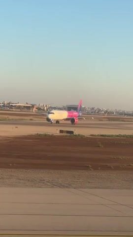 A Wizz Air airplane is visible on a runway. The plane is white with pink accents on the tail and engines. It appears to be either taxiing or preparing for takeoff. The sky is clear and blue, and the sun is shining. The background shows airport buildings and other distant aircraft. The foreground consists of the airport tarmac and some brown vegetation.