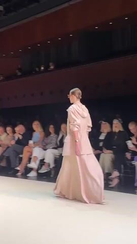 A fashion show is taking place in a darkened auditorium with tiered seating. A female model in a light pink hooded dress is walking down a runway, her back to the camera. Spectators are seated in the background, illuminated by stage lighting. The audience appears engaged, with some individuals looking at the model and others at their phones. The overall atmosphere suggests a professional fashion event.