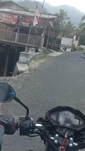 This is a first-person perspective video of someone riding a motorcycle. The rider is wearing a black helmet and a blue jacket. The motorcycle has a black mirror on the right side. The video shows a rural road with trees and vegetation on either side. There is a construction site visible with building materials and workers. The road appears to be unpaved in some areas. The video is shaky, indicating movement.