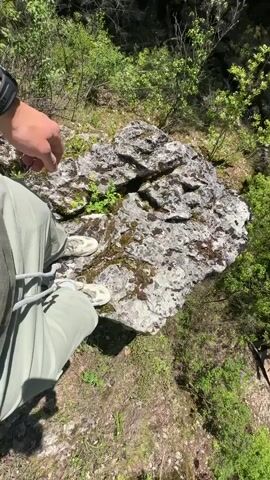 The video shows a person standing on a rocky ledge overlooking a green forested canyon. The person is wearing light green pants and light-colored shoes. They appear to be near the edge of a cliff. The camera angle is initially downward, showing the rock the person is standing on. It then slowly pans down to reveal the canyon below. There is a small pond or body of water in the canyon, with two wooden bridges leading to it. Lush green vegetation surrounds the water.  At the end of the pan, an archway of rock is visible, forming a natural bridge or cave.