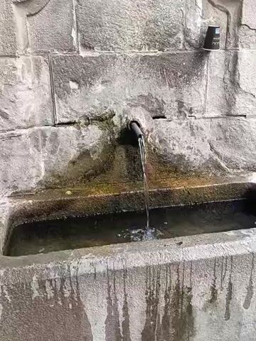 A stone fountain is seen with water flowing from a pipe into a stone basin. The stone wall behind the fountain has a rough texture. There is a disposable cup on the upper right of the frame. The water in the basin is disturbed by the flow from the pipe. The basin itself appears to be made of stone and has streaks of discoloration running down its sides. The scene appears to be outdoors.