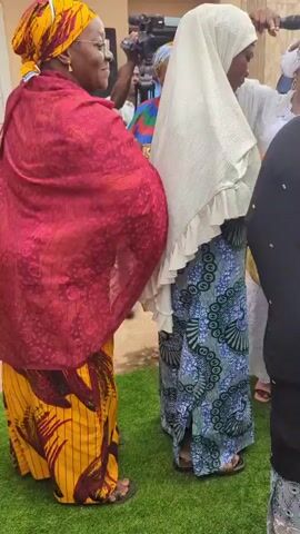 Several women in traditional attire are gathered outdoors on a green mat or carpet. The camera moves, following three women in the foreground. On the left, a woman wears a yellow and brown patterned dress with a red and black patterned shawl and a gold headwrap. In the center, a woman wears a white ruffled hijab and a long blue and black patterned skirt, carrying a yellow bag and a black bowl. A third woman, partially visible on the right, wears a black shawl over a patterned skirt and carries a black handbag. Other women are visible in the background, some smiling and clapping, wearing various colorful garments and head coverings. Tent structures are visible in the distance. The women in the foreground appear to be moving or dancing to music.