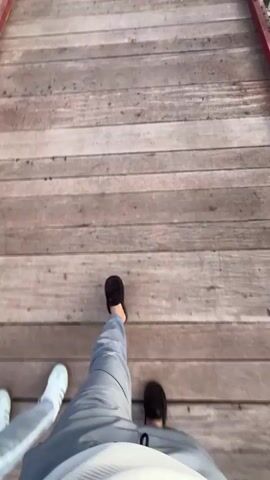 The video shows a person walking across a long, wooden suspension bridge. The bridge has white railings and red edges and is suspended over a body of water. In the background, there are mountains and a blue sky with clouds. The person is wearing gray pants and black shoes, and the camera is positioned at a first-person perspective, showing their legs as they walk.