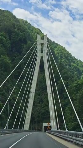 A car is driving on a bridge. The bridge has two large concrete towers with cables extending from the towers. The towers and cables are the focal point.  The bridge leads to a tunnel entrance. The surrounding environment features a lush green mountainside on either side of the bridge. The sky is partly cloudy. The overall scene is shot from the perspective of the car's dashboard.