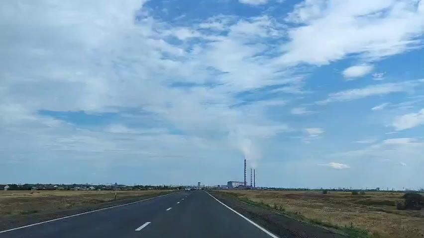 The video shows a view from a vehicle traveling on a highway under a partly cloudy blue sky. In the distance, a large industrial complex with several tall smokestacks emits plumes of white smoke. The surrounding landscape consists of dry, grassy fields with scattered low-rise buildings and vegetation along the left side of the road. Several vehicles are visible on the highway, driving in the same direction as the camera. The road has visible lane markings.