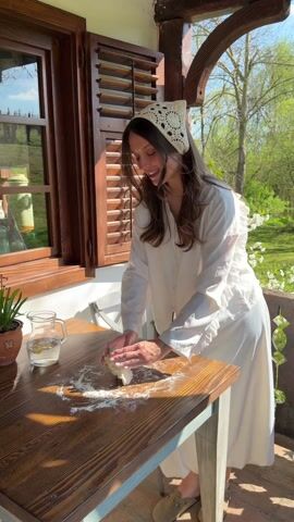 A woman is kneading dough on a wooden table outside on a porch. She is wearing a white, long-sleeved dress with ruffles, a white crocheted headscarf, and tan slippers. Flour is scattered on the table. A glass pitcher with water and a lemon slice, and a potted plant are visible on the table. The table is situated in front of a window with wooden shutters. Outside the window, trees with white blossoms are visible. She looks up and smiles.