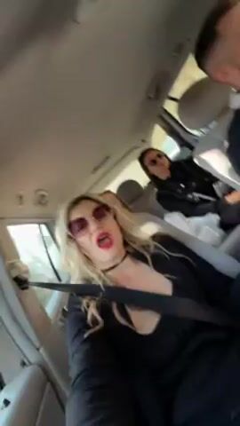 The camera is pointed forward inside a moving car. The interior is gray, including the car seats. Seated in the front passenger seat is a woman with blonde hair, wearing a black choker, a black low-cut top and blazer, and gold-rimmed sunglasses. She has bright red lipstick and is lip-syncing to music. She is wearing a seatbelt. Two other women are visible in the back seat, both wearing sunglasses and dark clothing. A man is driving the car, and his face is partially visible. Trees and sunlight can be seen through the windows.