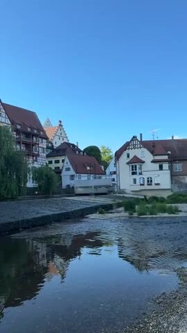 The video shows a calm river scene with buildings reflected in the water. The buildings are a mix of white and red-roofed structures with some half-timbered designs. A willow tree stands on the riverbank. The sky is clear and blue. The river is shallow, with visible stones at the bottom.