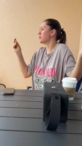 A woman with dark hair tied in a ponytail is seated at a dark gray outdoor table on what appears to be a balcony or patio in Al Fiqait, United Arab Emirates. She is wearing a light gray t-shirt with "ADVENTURE" printed in pink, along with an eagle graphic and "TRUE ATTITUDE" underneath. She has a small green earring in her left ear.

The woman is actively moving her hands and upper body, her facial expression animated and joyful, suggesting she is dancing or singing along to music. A black portable speaker is on the table in front of her. To her right, there is a white paper cup, likely from Starbucks, and a clear glass with a small amount of liquid. A smartphone is partially visible on the table to her left. The background features a light beige wall and a striped chair cushion. The scene is brightly lit, indicating it is daytime.