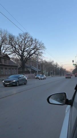 The video depicts a view from inside a car driving down a road. The road has tram tracks running down the center. Other cars are visible ahead. On the left side of the road, there is a wooden building and a large, bare tree. Traffic lights indicate a green light. The sky is clear and blue.