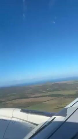 The video shows an aerial view from an airplane window. The sky is a clear, bright blue. Below, a landscape of varied terrain is visible, including flat areas and a mountain range in the distance along the coast. The wing of the airplane is visible in the foreground, with its white color contrasting with the blue sky and the landscape below.