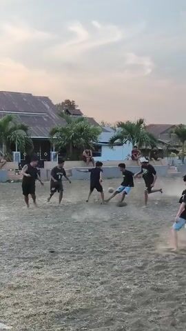 A group of young people are playing soccer on a dirt field. The sun is setting, casting a warm glow over the scene. In the background, there are houses and palm trees. The players are wearing casual clothing, and they are kicking the ball back and forth. One player is in the air, attempting to kick the ball. The game appears to be friendly and informal.