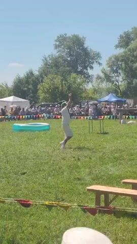 A person in a white outfit and a green hat is in a grassy field, facing away from the camera. The person is holding a white object, possibly a flag or a cloth, and appears to be throwing it. They are standing near a tall, thin pole. In the background, there is a crowd of people sitting and standing, some under tents. There is also a small inflatable pool to the left of the frame and a large inflatable slide in the distance to the right. The sky is clear and blue, and there are trees in the background. Wooden benches are visible in the foreground.