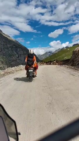 A first-person perspective video shows a dirt road winding through a mountainous landscape under a cloudy blue sky. A person wearing a white helmet and a backpack is riding a motorcycle with another person on the back. The rider is wearing an orange shirt and the passenger is wearing red pants. The motorcycle is moving away from the camera. The mountains on either side of the road are rocky and covered in patches of green vegetation. The road appears to be uneven and is made of dirt and gravel.