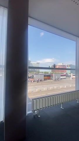 The video presents a short, indoor pan of what seems to be a modern meeting room. The room features large windows offering an exterior view of a street with people, vehicles, and buildings under a blue sky. Inside, there's a contemporary chair, a white conference table with a glass of water and a plate, and a coat rack. The interior is characterized by a neutral color palette of gray and white with a grid-patterned ceiling. The lighting appears to be a mix of natural light from the windows and overhead lighting.