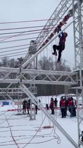 The video shows a person participating in a winter obstacle course. The individual, wearing a helmet and safety harness, is suspended from a metal structure and is maneuvering along a horizontal beam using ropes and their body weight. Snow covers the ground, and trees are visible in the background. Several people stand below, observing the participant. The course appears to be constructed from metal trusses and ropes. Light snow is falling.