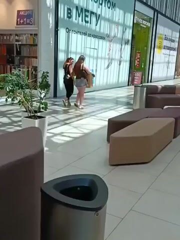 Two women are walking through a brightly lit, modern shopping mall. The woman on the left is wearing a black tank top and black leggings, and the woman on the right is wearing a black tank top and a short denim skirt. Both women are carrying shopping bags and appear to be walking towards the right side of the frame. Behind them, a large advertisement banner is visible with text in Russian. The mall interior features sleek, modern furniture, including a curved seating area in the foreground, and potted plants. The floor is tiled and reflects the overhead lighting. The overall impression is one of a typical shopping environment on a bright day.