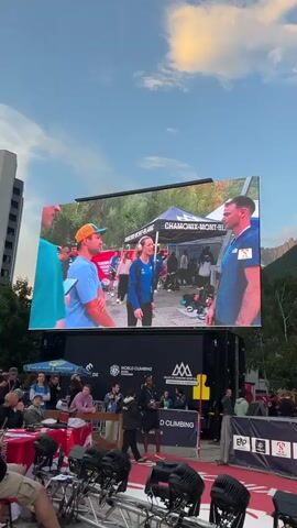 The video opens with a large outdoor LED screen displaying an event. The screen shows a man in a blue shirt with an orange hat and a man in a teal shirt. They appear to be in an outdoor setting with tents and other people in the background. Below the screen, there is a stage area with several people standing. A banner on the stage reads "WORLD CLIMBING CHAMONIX". There are also several rows of chairs and lighting equipment visible in the foreground. The sky is cloudy, and mountains are visible in the distance. The camera pans slightly to the left, revealing more of the crowd and the event setup. The screen then transitions to a white screen with some blurred red shapes.