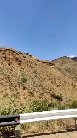 The video captures a view from a moving vehicle, looking out at a hilly, arid landscape under a clear blue sky with scattered clouds. A white guardrail with black stripes runs along the side of the road in the foreground. A tall, thin pole with a triangular sign is visible on the left. The terrain is characterized by dry, scrubby vegetation covering gentle slopes and steeper, rocky inclines. In the distance, larger mountains are visible. The camera's perspective shifts slightly, offering a panoramic glimpse of the natural scenery.