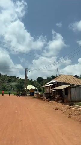 The video begins with a view of a dirt road in what appears to be a rural or developing area. The road is reddish-brown and a bit dusty. On the right side of the road, there are a few structures that look like small shops or homes, made of corrugated metal and other simple materials, with thatched or tin roofs. A power pole with wires runs across the scene. In the background, there are green hills with rocky outcrops and trees. The sky is blue with large, fluffy white clouds.

As the video progresses, the perspective shifts. We see people walking on the road. There are a few vehicles, including what looks like a white sedan and a three-wheeled auto-rickshaw (also known as a tuk-tuk). Some people are standing around, possibly interacting or resting. One scene shows a few individuals near a small building with a yellow awning. Another shot captures a tuk-tuk driving away from the camera. The environment remains consistent with hills, vegetation, and the dirt road. The overall impression is of a sunny day in a community with basic infrastructure.