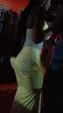 A person wearing a light-colored, form-fitting dress dances in a dark outdoor setting, likely a party or gathering. The camera focuses on the individual's back and side profile as they move to music. Other indistinct figures are present but blurred in the background. The ground is dark and potentially uneven, with hints of red light.  The video quality is grainy and the lighting is poor, making it hard to discern specific features.