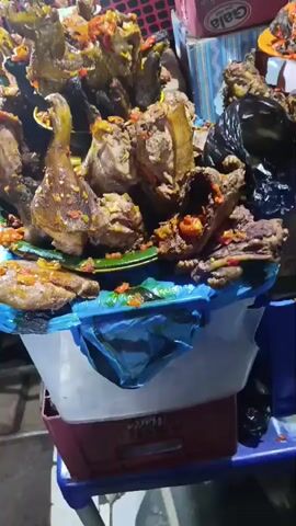 The video captures a close-up view of a food stall, likely in an outdoor market setting, at night. The primary focus is on a large display of cooked meat, predominantly appearing to be poultry (chicken or turkey) and possibly other cuts, piled high on various platters and in containers.

The meat is heavily seasoned with a vibrant mix of red and yellow spices or peppers, giving it an appetizing and colorful appearance. Some pieces are whole or half-carcasses, while others are cut into smaller portions, ready for sale. Below the main display, more meat is visible, including what looks like a flattened piece of chicken or fish.

The display is set up on what appears to be stacked plastic crates or containers, covered with blue plastic sheeting. In the background, to the right, a red box with "Gala Classic Sausage Roll" printed on it is visible, suggesting other items are also sold. Another container at the bottom partially shows the word "Malta". The general ambiance is dimly lit, typical of a night market, with artificial lights illuminating the food. No people are explicitly visible, but the arrangement clearly indicates an active food business. The background is a dark area with a red, patterned mesh or fabric.