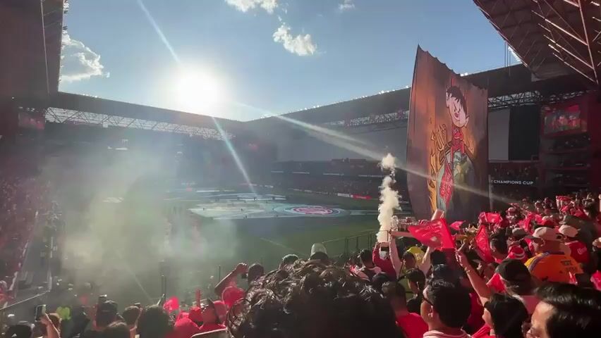 A wide, high-angle shot shows a packed stadium filled with fans primarily wearing red, some waving red flags. The bright sun shines overhead. On the right side of the frame, a large banner depicts a cartoonish figure. In front of the crowd, smoke fills the air, possibly from flares or pyrotechnics, partially obscuring the field. In the foreground, a person is holding up a tray full of red plastic cups, and several people are filming with their phones. In the background, performers are on the field, and screens display game information and advertising. The stadium architecture is visible, with a red roof structure. The scene is likely a sporting event, possibly soccer, given the "Champions Cup" signage.