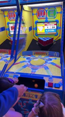 The video features a man and two young girls playing a "DUNK IT!" arcade game in what appears to be an entertainment center or arcade in Columbus, United States. The game machine is brightly colored in blue, yellow, and red.

The man, wearing a light-colored top with blue sleeves, helps one of the girls retrieve an orange ping-pong ball from a compartment at the base of the machine. One girl, with long dark hair and wearing a yellow shirt, stands in front of the machine, actively throwing a ball towards the game's target. Another younger girl, with dark hair and a bow, sits below, watching the action.

The game's digital screen shows "YOUR SCORE 1," "BALL 1," and the number "24," possibly indicating a remaining ball count or timer. Text on the screen encourages players to "TRY TO HIT ALL 10 CUPS!" Above the main screen, a bonus display outlines ticket rewards for different numbers of cups hit: "0-3 = 10," "4-6 = 14," "7-9 = 20," and "10 = JACKPOT!" It also advertises "10 TICKETS EVERY BALL IN LAST 10 CUPS HAVE BEEN HIT!" The game offers options for "HEAD-TO-HEAD COMPETITION" and "2-PLAYER LINKED GAME PLAY." The children appear engaged and focused on the game.