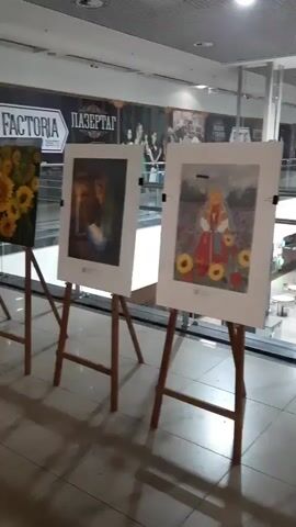 A video displays a row of artwork displayed on easels in what appears to be a mall. The artwork consists of framed prints or paintings, each on its own wooden easel. The first piece of art from the left is dark and appears to depict a solitary figure in a dimly lit setting. The next piece shows a colorful depiction of a person in a red outfit surrounded by sunflowers. Following that, a landscape painting with trees and a mountainous background is visible. The subsequent artwork features a portrait of a person with flowing hair against a backdrop of blues and oranges. Another framed piece shows two images of dogs lying down. The next artwork in the sequence is a busy scene with many small figures. This is followed by a painting of a building or structure. The last visible artwork is a portrait of a woman's face with red elements. The background suggests a modern indoor environment with glass partitions, escalators, and other shops. The lighting is artificial, coming from overhead fixtures. The floor is tiled. No people are actively engaged with the artwork, but there are glimpses of activity in the background through the glass.