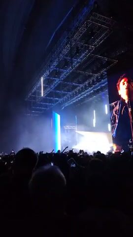 The video shows a concert from the perspective of someone in the audience. The crowd is silhouetted against a bright stage with various light displays including vertical bars of light and smoke. A large screen on the right side of the stage shows a man singing. The audience has their hands up and some are recording the performance with their phones. An intricate metal structure is visible above the stage.