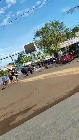 The video shows an outdoor scene with a dirt road and a few buildings in the background. There are a group of people standing in a line on the road, some of whom appear to be tourists. There are also several vehicles present, including cars, motorcycles, and a bus. The sky is blue with some clouds, and the overall lighting suggests it is daytime. The area appears to be a rural or semi-urban location.