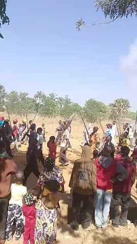 The video depicts a crowd of people gathered in an outdoor setting, possibly a field or open area with trees and shrubs in the background. The sky is clear and blue. Many in the crowd are holding what appear to be rifles or long guns, raised in the air. Some individuals are dressed in casual clothing, while others wear more traditional attire. Several children are present in the foreground. Gunfire is audible, and smoke is visible near the center of the frame, suggesting the firing of weapons. A flag with white, green, and black colors is also visible.