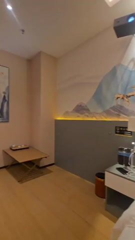 A virtual tour of a hotel room. The room has wood floors and pale pink walls. In the first shot, a low wooden table with tea service is positioned in front of a landscape painting. The next shots show a bathroom with green tile and a vanity, followed by a luggage rack with a rose gold suitcase. Finally, a large projector screen is displayed on the wall with a shelf underneath holding water bottles and an orange box.