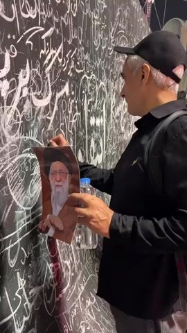 A man wearing a black shirt and a black baseball cap is writing on a black wall covered in white Arabic calligraphy. He is holding a photograph of an elderly man with a white beard and glasses in his left hand, and with his right hand, he is using a piece of chalk to write on the wall. A water bottle is visible on the wall behind his left hand. The man is facing the wall and looking down at his writing. In the background, other people can be seen, blurred and out of focus.