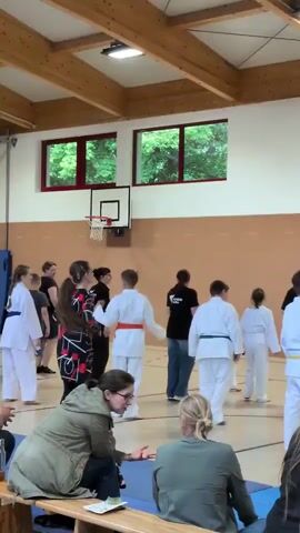 The video captures an indoor scene, likely a gymnasium, with a group of people, mostly children and teenagers, dressed in martial arts uniforms, primarily white gi with colored belts. They appear to be engaged in a training session or demonstration. In the foreground, two adults are seated, observing the activity. The background reveals a typical gymnasium setup with wooden ceiling beams, basketball hoop, and windows showing greenery outside. The participants are arranged in a semi-circle, facing a central figure or area where instruction seems to be taking place. Some individuals are performing movements, while others are standing and listening. The atmosphere appears to be one of focused activity and instruction.