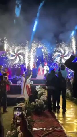 The video shows a wedding celebration with fireworks. A bride in a red and white dress and a groom in white stand on a stage, surrounded by people. Bright lights and exploding fireworks illuminate the scene, creating a celebratory atmosphere. Guests record the event with their phones. The background features floral decorations and other decorative elements.