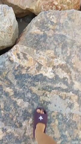 The video begins with a close-up of large, rough-textured rocks. A person's right foot wearing a dark purple sandal with white text is visible, stepping onto one of the rocks. The camera then pans upwards to show a pathway made of similar large rocks stretching into the distance, forming a breakwater. The sea is visible on both sides of the breakwater, with moderate waves. The sky above is blue with some scattered clouds. The video continues to move along the rock pathway, showing the expanse of the sea and sky.