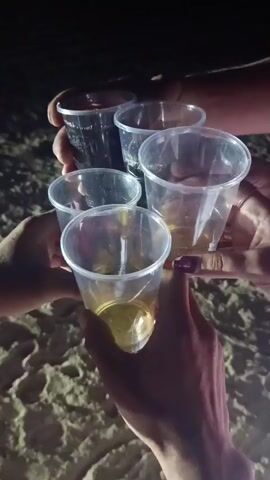 The video begins with a close-up shot of several hands holding up plastic cups filled with liquid. The cups are held together in a toast. The scene is dark, suggesting it is nighttime. The background is blurred but appears to be sand, indicating the setting might be a beach. As the video progresses, the camera pans up and away from the cups, revealing a dark sky with distant lights and what appear to be fireworks. The fireworks are red and burst in a bow-tie shape. The overall mood is celebratory.